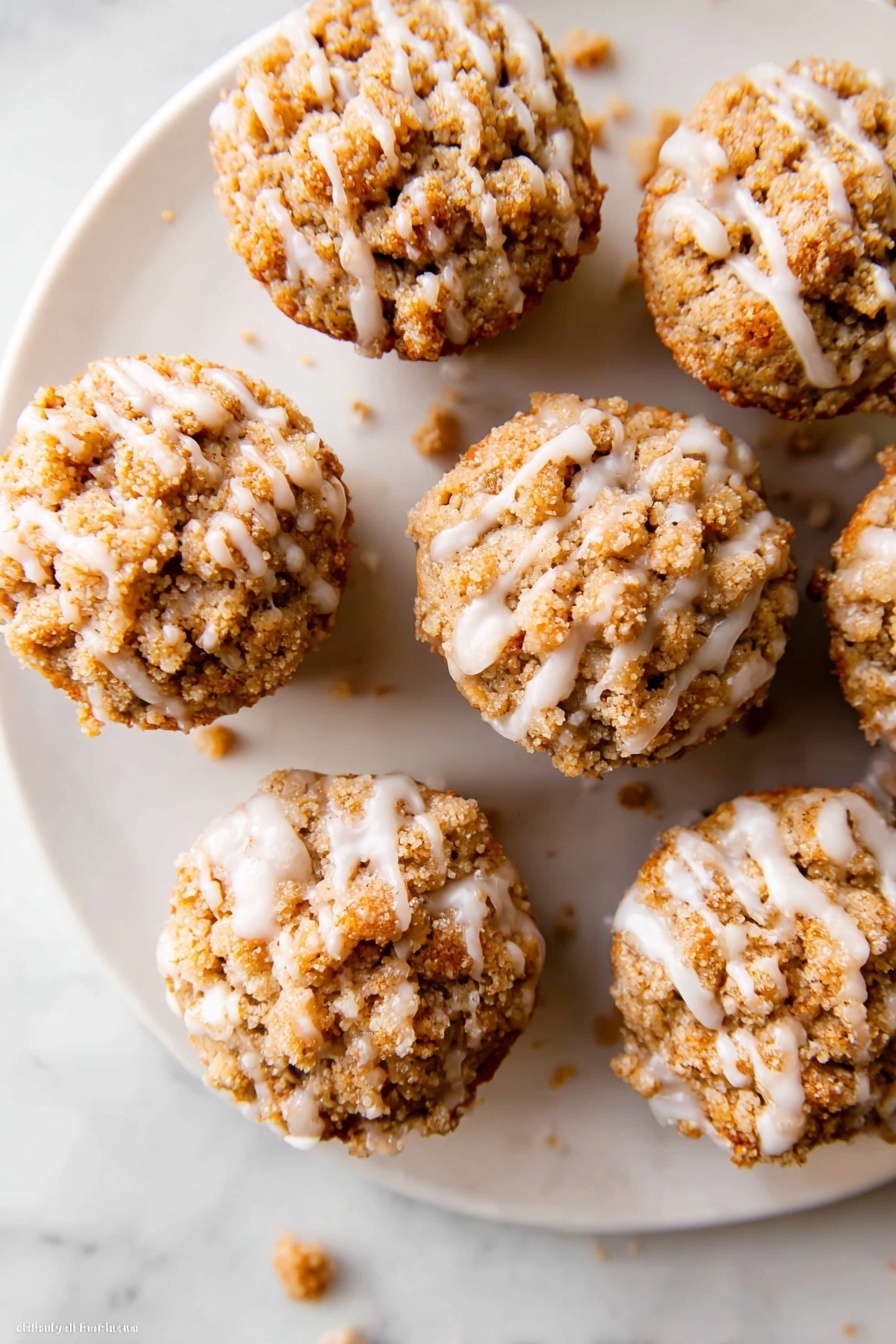 Pumpkin Crumb Muffins with Maple Icing, Pumpkin Muffins, Fall Muffin Recipes, Moist Pumpkin Muffins, Easy Pumpkin Muffin Recipe - The image shows a white plate with six small muffins arranged in a loose circle on a white marbled surface. Each muffin has a crumbly, light brown streusel topping that covers the whole top and looks crunchy. A thin layer of white icing is drizzled unevenly over the muffins, creating shiny lines that drip slightly onto the plate. A few crumbs are scattered around the plate and on the marbled surface. The muffins look moist and textured with a mix of crumbly and smooth parts. Photo taken with an iphone --ar 2:3 --v 7