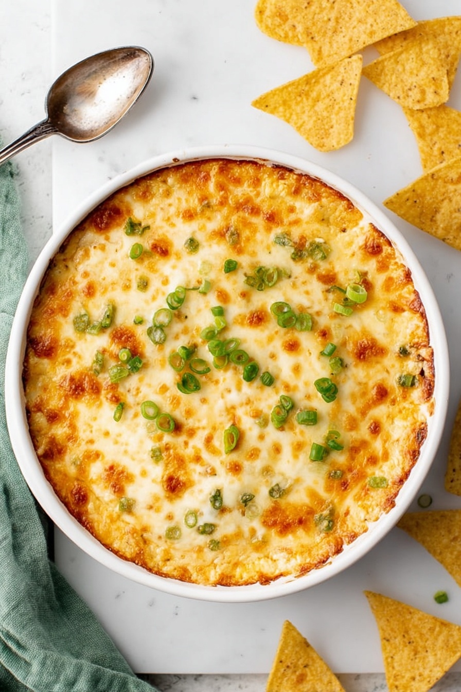 Crab Rangoon Dip with Crispy Wonton Chips, crab rangoon dip, creamy crab dip, appetizer with wonton chips, cheesy crab dip - A round white baking dish filled with a golden, bubbly cheese layer on top, showing patches of light brown where the cheese is slightly toasted, scattered with small rings of bright green chopped scallions. The entire dish sits on a white marbled surface with a vintage silver spoon to the side, broken and whole triangular yellow tortilla chips nearby, and a soft green cloth partly visible at the edge. Photo taken with an iphone --ar 2:3 --v 7