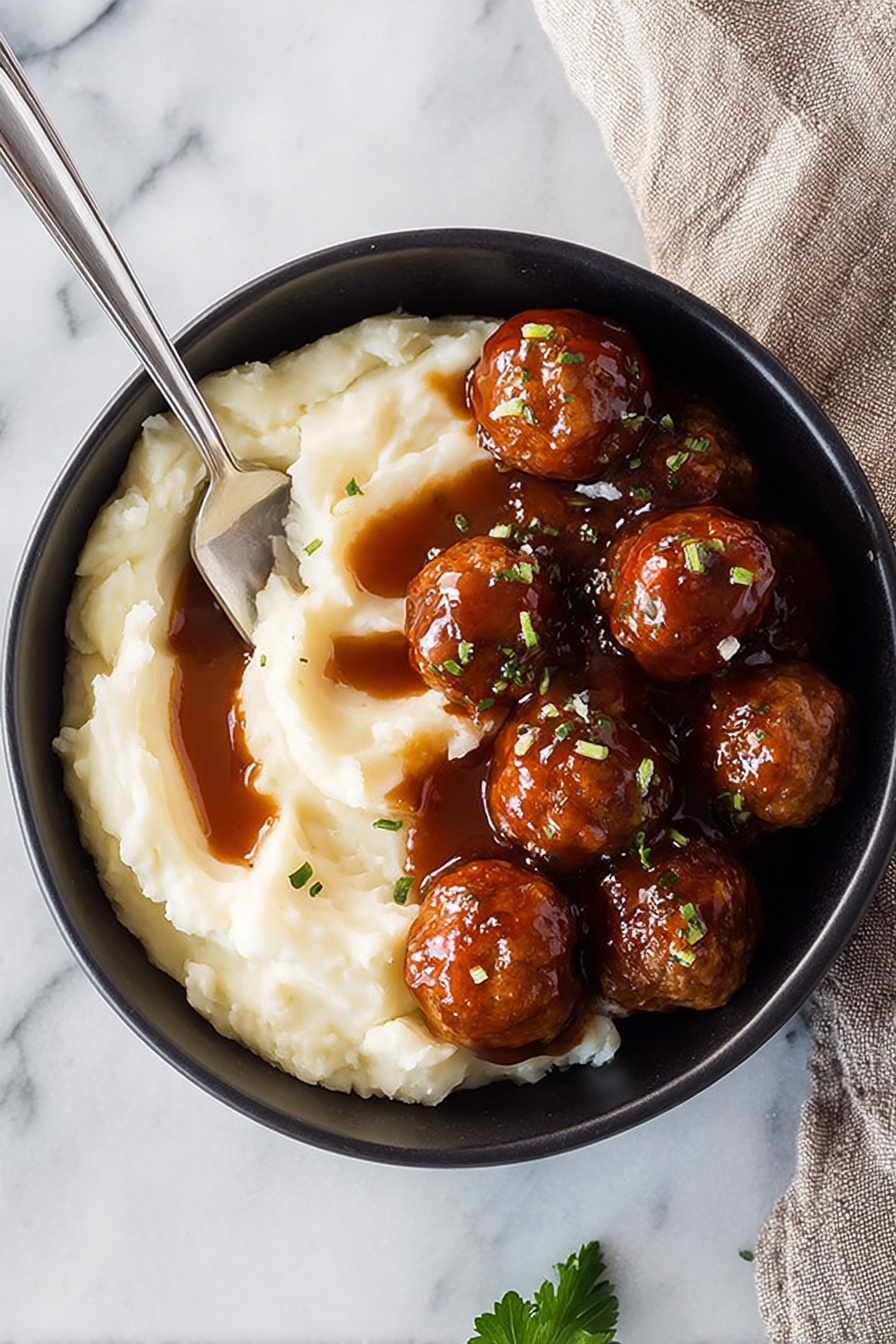 Bourbon Slow Cooker Meatballs, easy meatball appetizers, slow cooker meatball recipes, party meatballs with bourbon, flavorful meatball dinner - A black bowl on a white marbled surface holds a serving of mashed potatoes and meatballs. The mashed potatoes form a soft, creamy layer on one side, with a smooth texture and three streaks of dark brown gravy on top. On the other side, a cluster of round, shiny meatballs covered in thick brown sauce are garnished with small green herb pieces. A silver spoon rests partially sunk into the mashed potatoes, angled towards the bowl's edge. A small parsley leaf lies nearby on the surface. photo taken with an iphone --ar 2:3 --v 7