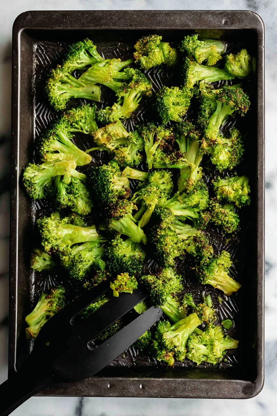 Oven Roasted Broccoli with Garlic and Paprika, roasted broccoli side dish, garlic roasted broccoli, smoky roasted broccoli, easy roasted broccoli recipe - A dark rectangular baking tray holds a single layer of cooked broccoli florets. The broccoli pieces are bright green with some slightly browned tips, showing a crispy texture. The florets vary in size and shape, spread unevenly across the tray. In the bottom right corner, a black spatula is lifting some broccoli. The tray rests on a white marbled surface. photo taken with an iphone --ar 2:3 --v 7