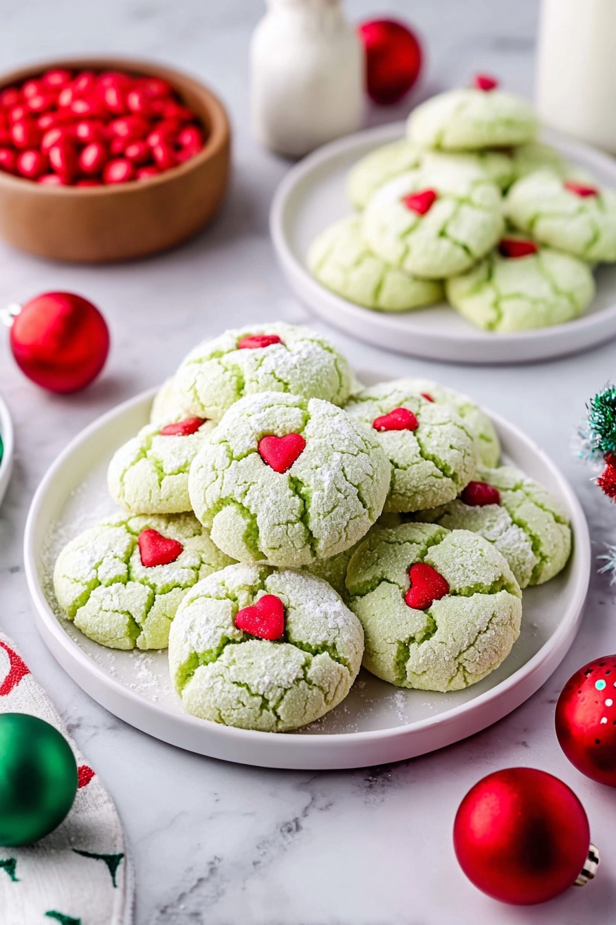 Green Peppermint Cake Mix Cookies, festive peppermint cookie recipe, easy holiday cookies, quick cake mix cookies, minty Christmas treats - A large pile of light green cookies with a soft cracked texture, each topped with a small red heart candy, sits on a white plate in the center. Two smaller white plates in the background hold more of the same cookies. The cookies are dusted lightly with powdered sugar. Around the plates are red and green round Christmas ornaments and a small wooden bowl filled with red candies on a white marbled surface. A white bottle is visible in the background. Photo taken with an iphone --ar 2:3 --v 7