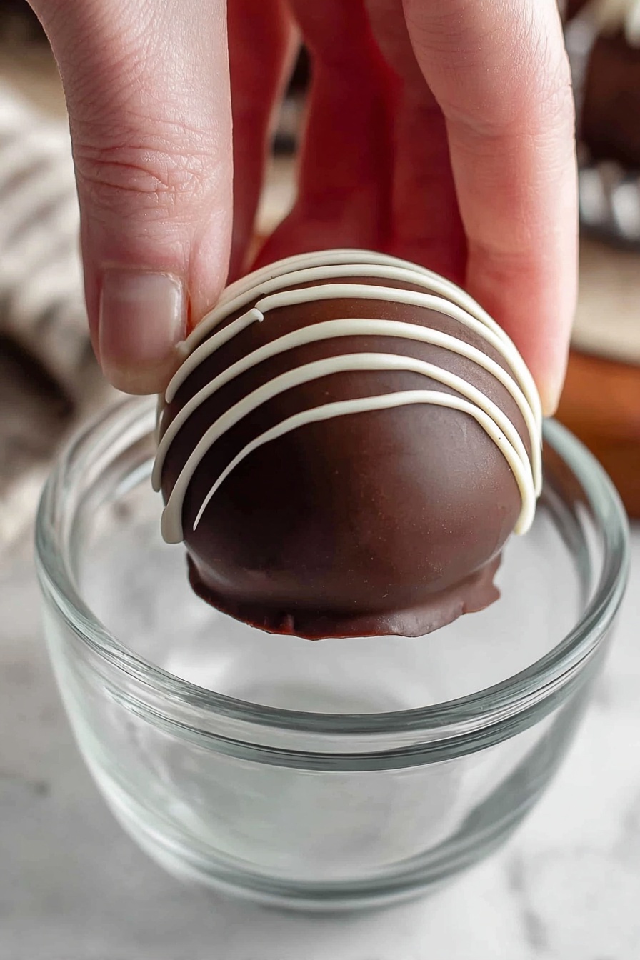 Homemade Hot Chocolate Bombs, hot chocolate bombs recipe, how to make hot chocolate bombs, DIY hot chocolate bombs, hot cocoa bombs at home - A woman's hand is holding a round dark chocolate ball with white thin lines of icing drizzled vertically on top. The chocolate ball has two layers, the outer smooth dark chocolate shell, and the white icing on the top. It is being held above a clear glass cup with a simple design. The background shows a white marbled textured surface. photo taken with an iphone --ar 2:3 --v 7