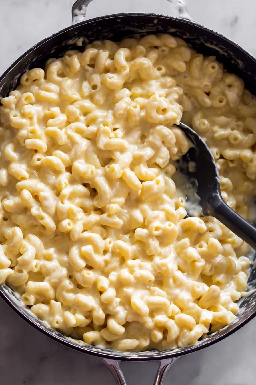 Creamy Baked Mac and Cheese, baked mac and cheese, cheesy baked pasta, comfort food recipes, homemade mac and cheese - The image shows a black pan filled with creamy macaroni and cheese. There is one thick layer of soft elbow pasta coated in smooth, light yellow cheese sauce. A black spoon is partly submerged in the macaroni, lifting a scoop on one side of the pan. The pan is set on a white marbled surface which is slightly visible around the edges. Photo taken with an iphone --ar 2:3 --v 7