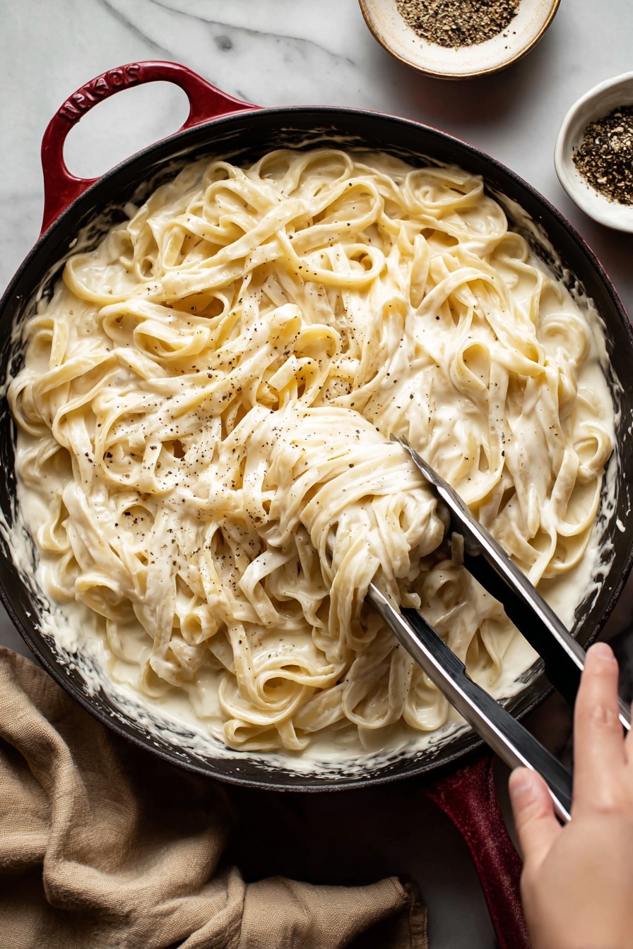 Vegan Alfredo Sauce with Cashews, vegan Alfredo sauce, dairy-free Alfredo sauce, cashew Alfredo sauce, plant-based Alfredo sauce - A close-up view of creamy fettuccine pasta served in a black pan with a red handle edge, the pasta is covered in a thick, smooth white sauce that looks rich and velvety; the long pasta strands are mixed well with the sauce and lightly sprinkled with black pepper; a woman's hand is holding metal tongs, lifting some pasta from the pan; in the background, there is a small white bowl with black pepper, and the whole setting is on a white marbled surface with a beige cloth nearby photo taken with an iphone --ar 2:3 --v 7