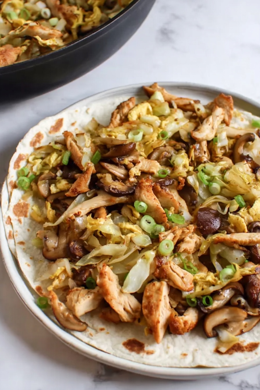 Moo Shu Chicken, easy Chinese stir-fry, quick dinner ideas, healthy Chinese recipes, veggie-packed stir-fry - This image shows a close-up of a black frying pan filled with a stir-fry dish. The dish has many pieces of brown, cooked chicken strips mixed with light yellow cabbage slices and orange-yellow egg strips. There are also some sliced mushrooms with a dark brown outer edge and light beige inside, scattered throughout. Small green onion pieces are sprinkled over the dish adding a splash of green color. The frying pan is set on a white marbled surface. photo taken with an iphone --ar 2:3 --v 7