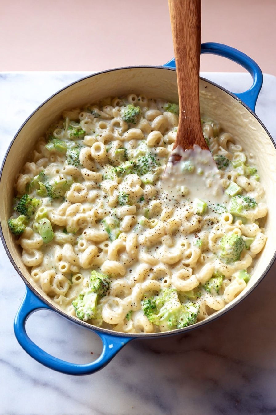 Creamy Broccoli Pasta with Cheesy Sauce, creamy broccoli pasta, cheesy pasta recipes, comforting broccoli pasta dish, easy cheesy pasta - A large white pot with blue handles is filled with small tube pasta and small green broccoli pieces mixed in a thick, creamy white sauce with visible black pepper specks, all stirred by a wooden spoon standing upright in the pot, placed on a white marbled surface with a soft pink background, photo taken with an iphone --ar 2:3 --v 7
