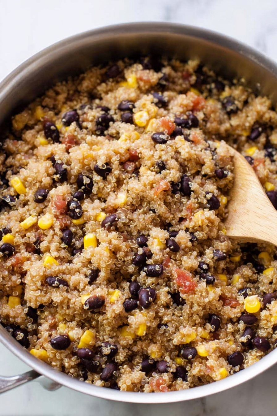 Easy Mexican Quinoa Bowl, Mexican quinoa bowl ingredients, quick healthy quinoa bowls, vegetarian Mexican quinoa meal, simple Mexican quinoa dinner - A silver pan filled with cooked quinoa mixed with black beans, yellow corn, and small pieces of red tomatoes. The quinoa grains are light brown and fluffy, evenly spread throughout the pan. The black beans are scattered, showing their dark smooth texture while the yellow corn adds bright pops of color. A wooden spoon rests inside the pan on the right side, partially covered by the quinoa mixture. The background is a white marbled surface. photo taken with an iphone --ar 2:3 --v 7