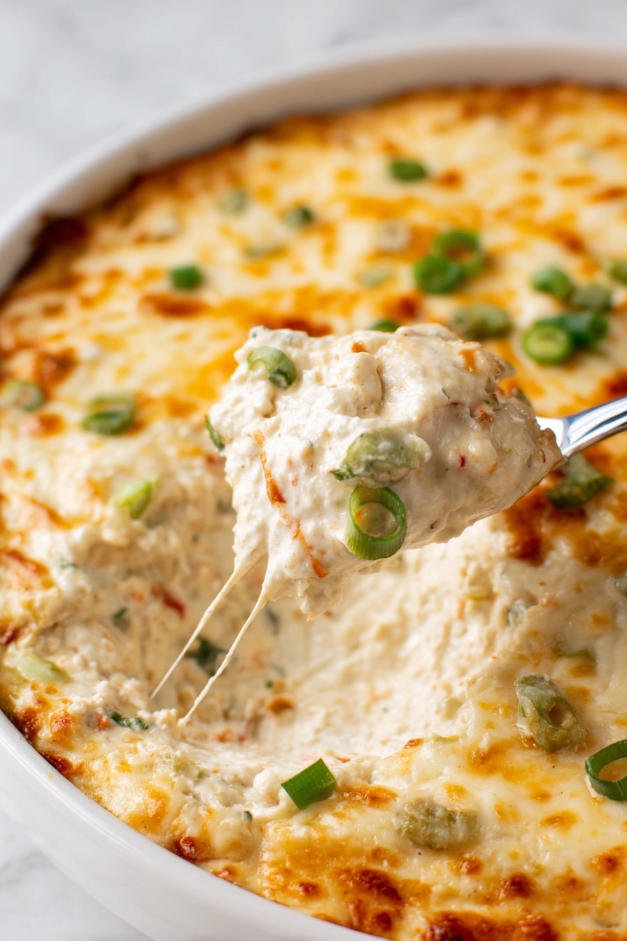 Crab Rangoon Dip with Crispy Wonton Chips, crab rangoon dip, creamy crab dip, appetizer with wonton chips, cheesy crab dip - A close-up of a white dish filled with a creamy dip that has a thick melted cheese layer on top. The cheese is golden with light brown spots and sprinkled with small green onion pieces. The creamy layer underneath is light beige with visible small bits of other ingredients, showing a rich and creamy texture. A spoon is lifting a scoop of the dip, revealing the thick inside. The dish is on a white marbled surface. photo taken with an iphone --ar 2:3 --v 7