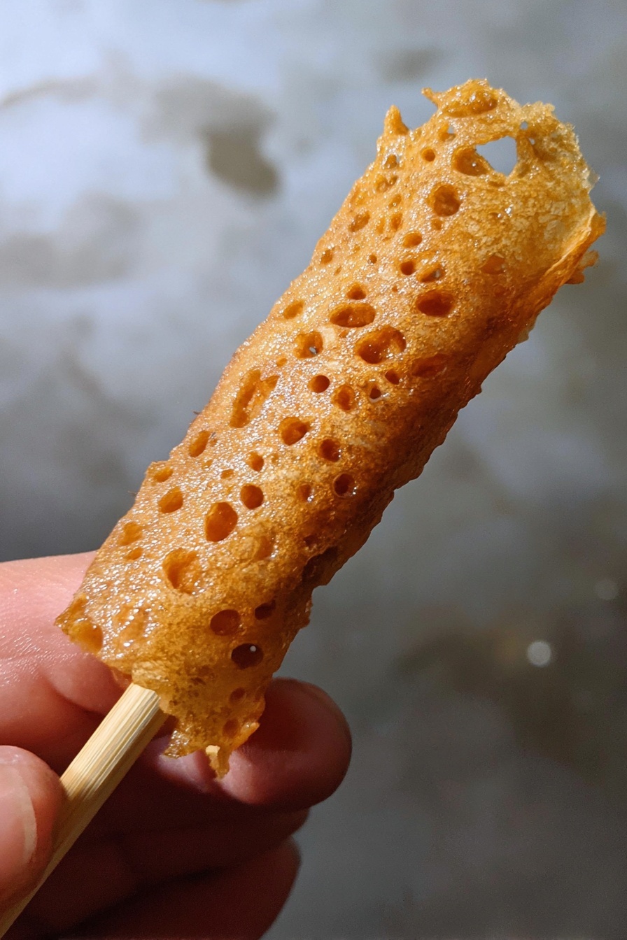 Brandy Snap Cookies, crispy caramel cookies, homemade brandy snaps, holiday cookie recipes, delicate cookie treats - A close-up image of a golden brown, thin, lace-like fried snack wrapped around a light wooden stick held by a woman's hand, with a textured, crispy surface that has small holes and a shiny finish, against a background with a white marbled texture. photo taken with an iphone --ar 2:3 --v 7