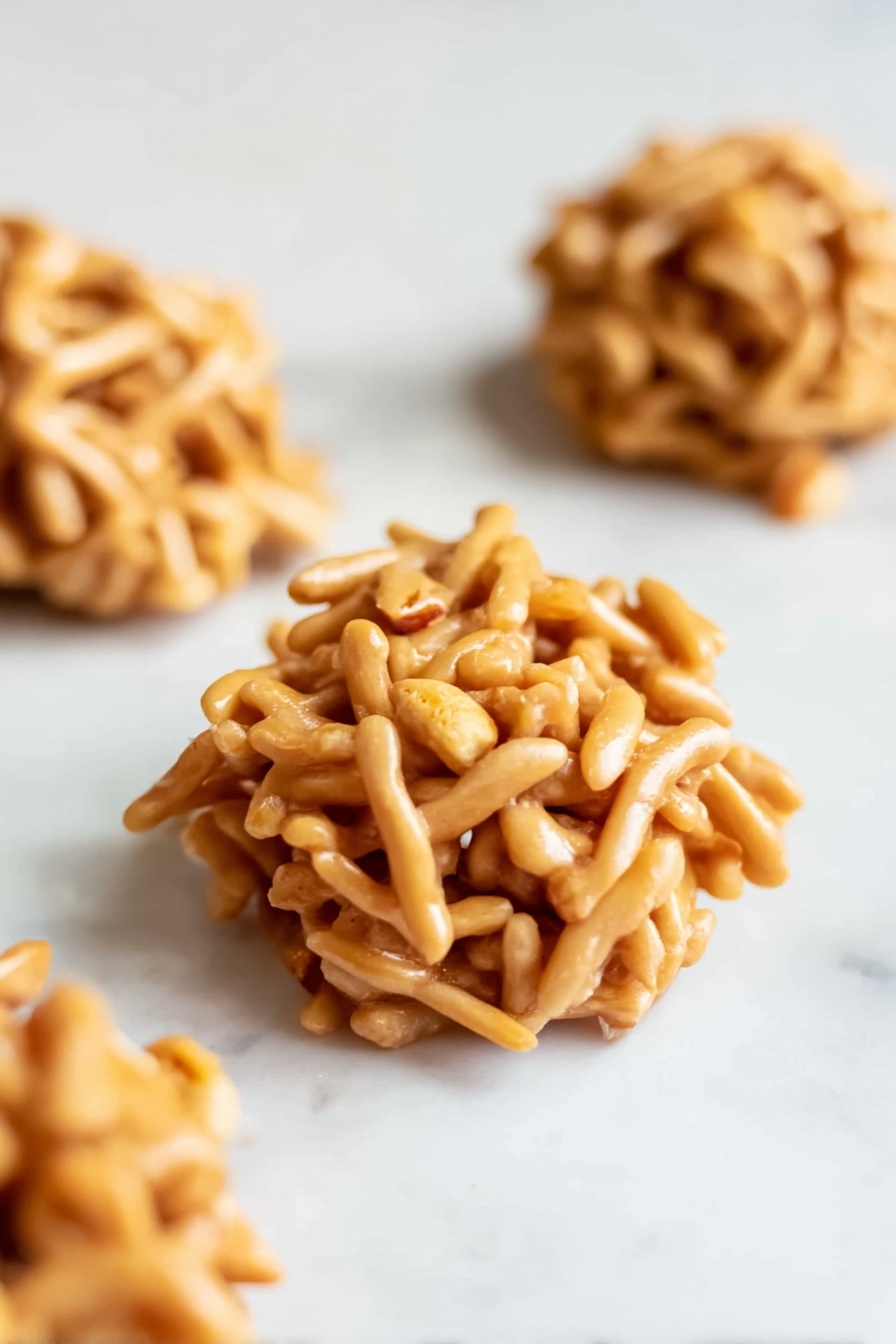 No Bake Haystack Cookies, easy no bake cookies, peanut butter chow mein cookies, no bake sweet treats, quick dessert recipes - The image shows small round clusters made of light brown, thin, stick-like pieces intertwined with small peanut bits visible in the mix. Each cluster is a single layered mound, with a rough and irregular texture, placed on a smooth white marbled surface. There are three clusters scattered, with one in clear focus at the front and the other two slightly blurred in the background. The sticks have a matte finish with subtle shadows giving depth and softness to the clusters photo taken with an iphone --ar 2:3 --v 7