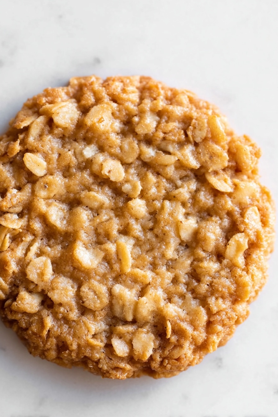 Lace Cookies with Brown Butter and Oats, crunchy lace cookies, nutty oat cookies, crispy brown butter cookies, delicate oat lace cookies - The image shows a close-up view of a single oatmeal cookie. The cookie is round and thick with an uneven surface made of baked oats. Its color is a warm golden brown with lighter patches of oat flakes throughout, giving it a rough texture. The cookie sits flat on a white marbled surface, and the edges are slightly raised and crispy-looking. There are no other objects or decorations in the frame, focusing all attention on the detailed texture of the oatmeal cookie. photo taken with an iphone --ar 2:3 --v 7