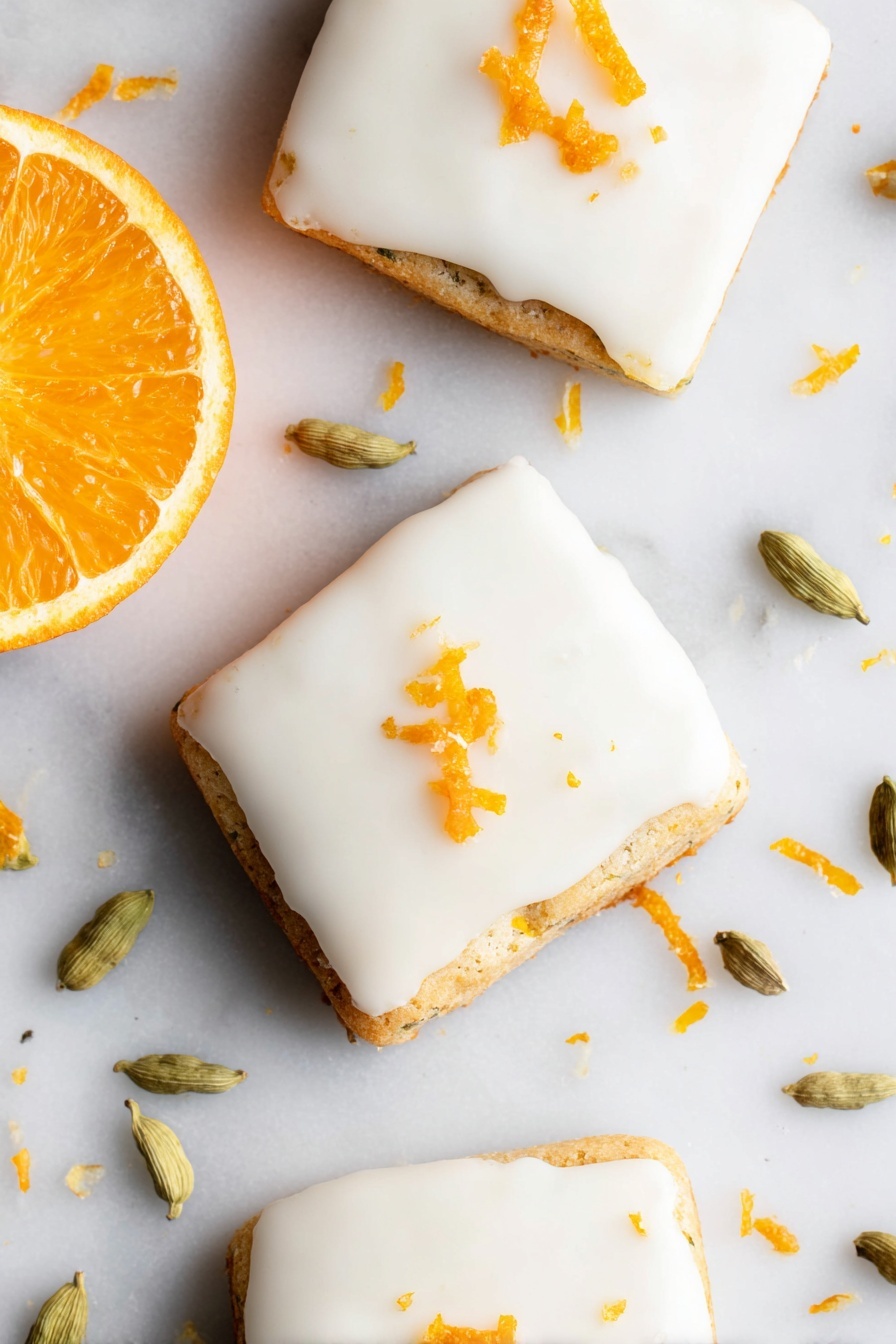 Cardamom Shortbread Cookies with Orange Glaze, citrus shortbread cookies, flavored holiday cookies, spiced butter cookies, orange glaze dessert - The image shows three square-shaped cookies with smooth white icing covering the top layer. Each cookie is slightly thick, with the base layer visible under the icing, showing a light golden-brown color and a slightly textured edge. Small pieces of orange zest are scattered on top of the icing, adding a bright orange color contrast. To the left side of the cookies, there is a fresh orange half with a vibrant, juicy orange color. Around the cookies on the white marbled surface, there are several light green cardamom pods and scattered bits of orange zest. The photo taken with an iphone --ar 2:3 --v 7