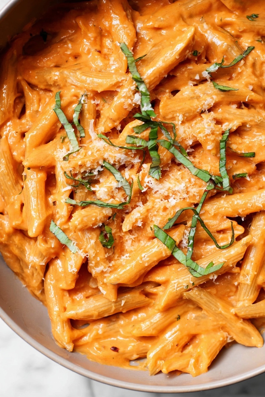 Creamy Penne alla Vodka, Penne alla Vodka, Vodka Pasta, Creamy Pasta Recipes, Easy Italian Pasta - The image shows a close-up of penne pasta covered in a thick, smooth orange sauce. The pasta is mixed well with the sauce and topped with thin green strips of fresh basil scattered across the surface. There is a light sprinkle of grated cheese that adds a slightly grainy texture on top. The background is a white marbled texture, making the colors of the pasta and sauce stand out clearly. photo taken with an iphone --ar 2:3 --v 7