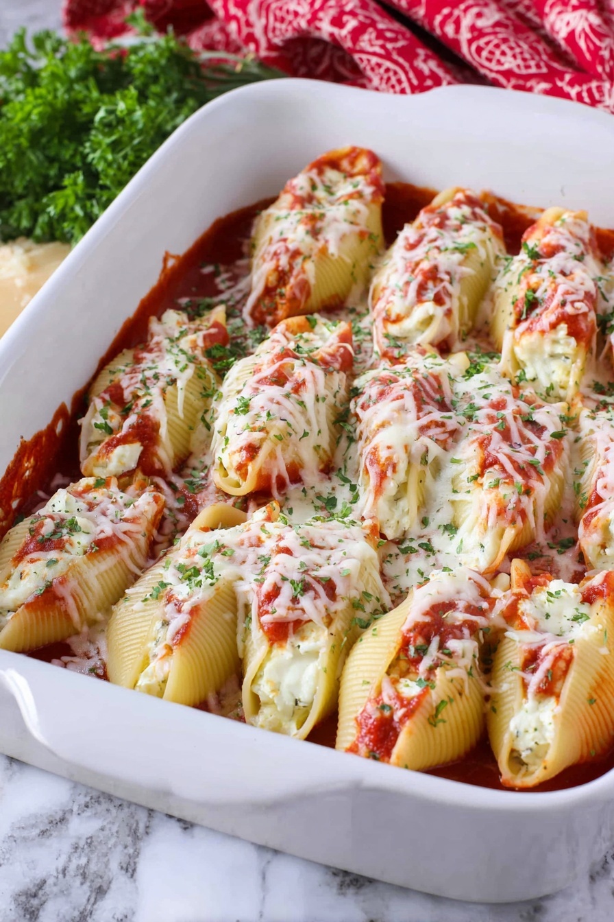 Cheesy Chicken Stuffed Shells, cheesy stuffed shells, chicken pasta bake, baked stuffed shells recipes, cheesy pasta shells - The image shows a white baking dish filled with two layers of large pasta shells. The bottom layer is covered with a red tomato sauce, and the shells are stuffed with white cheese inside, visible in some openings. The top layer consists of more pasta shells placed closely next to each other, covered with melted white cheese sprinkled with green herbs and bits of red tomato sauce. The dish is placed on a white marbled surface, with some green herbs and a red patterned cloth in the background. Photo taken with an iphone --ar 2:3 --v 7