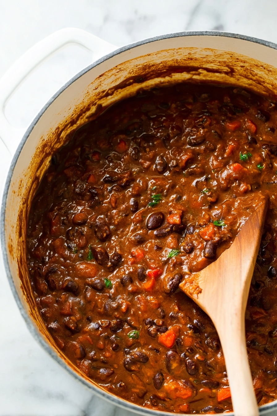 Vegetarian Chili, hearty vegetarian chili, easy vegetarian chili recipe, healthy chili recipe, flavorful vegetarian chili - A close-up view of a thick stew-like dish inside a large white pot with a white marbled surface underneath. The dish has a rich brown color with visible layers of ingredients, including dark brown beans, small red chunks, and some green herb bits mixed throughout. A wooden spoon is partly dipped in the stew on the right side, showing the texture which is soft and slightly chunky with a glossy finish. The sides of the pot have some sauce splashes and the scene looks warm and hearty. Photo taken with an iphone --ar 2:3 --v 7
