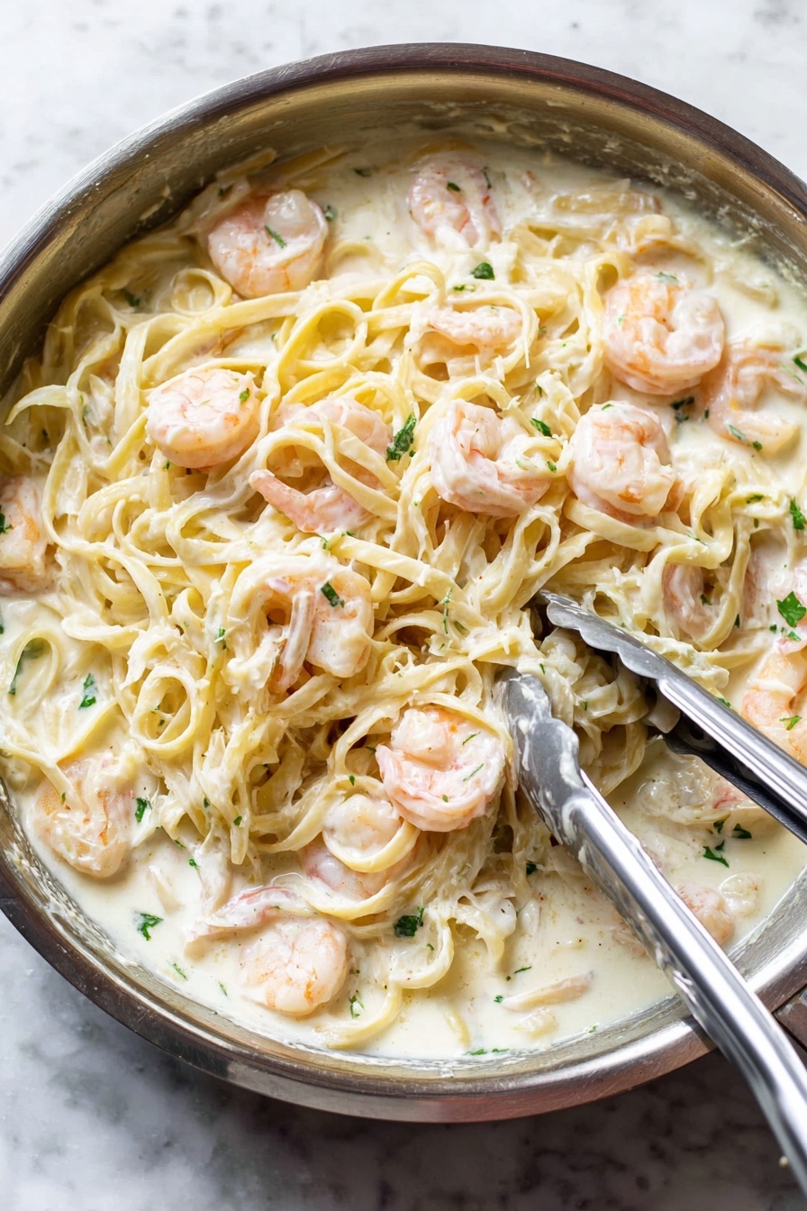 Shrimp Alfredo, easy shrimp pasta, creamy seafood pasta, quick shrimp Alfredo, simple Italian shrimp dish - A close-up view of a silver pan filled with creamy white sauce pasta mixed with pink shrimp. The pasta, light yellow and smooth, is covered in a thick white sauce and evenly spread throughout the pan. Plump shrimp are scattered on top and mixed in, their soft pink color contrasting with the white sauce. Small green herb pieces are sprinkled over the dish for a fresh touch. A pair of metal tongs and a spoon are resting on the pasta, indicating the dish is being stirred or served. The pan sits on a white marbled surface. photo taken with an iphone --ar 2:3 --v 7