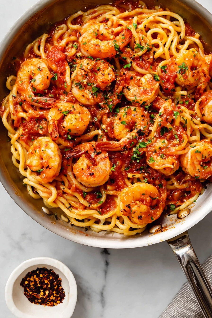Shrimp Fra Diavolo Pasta, spicy seafood pasta, quick Italian dinner, easy shrimp pasta recipe, flavorful seafood pasta - The image shows a black pan full of curly pasta mixed with cooked shrimp. The pasta is light yellow with a slightly wavy texture, while the shrimp are orange with reddish highlights, laid on top and mixed in all around. There is a red tomato sauce spread unevenly on the pasta and shrimp, and small bits of green herbs sprinkled across the dish. The pan is on a white marbled surface with a small white bowl of red chili flakes beside it at the bottom left. The metal handle of the pan extends out to the right side. Photo taken with an iphone --ar 2:3 --v 7
