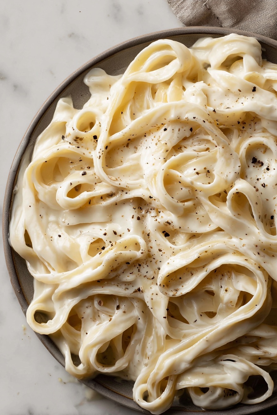 Vegan Alfredo Sauce with Cashews, vegan Alfredo sauce, dairy-free Alfredo sauce, cashew Alfredo sauce, plant-based Alfredo sauce - This image shows a close-up view of wide fettuccine noodles covered in a thick, creamy white sauce. The sauce looks smooth and rich, evenly coating the noodles. Light brown small specks of black pepper are scattered across the pasta, adding texture and contrast. The dish fills the frame, with noodles tangled and folded over each other in layers. The background is a white marbled surface. photo taken with an iphone --ar 2:3 --v 7