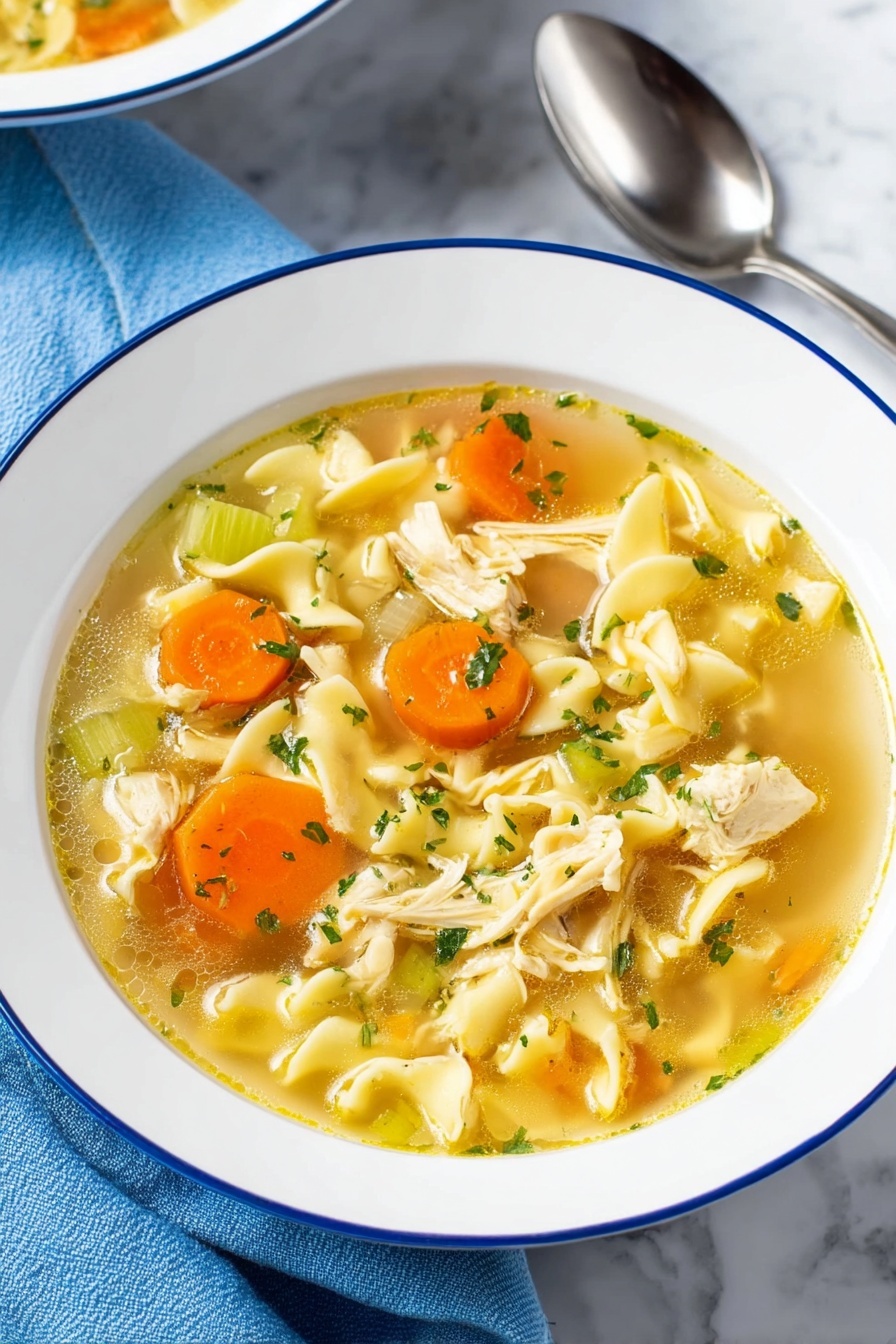 Vegan Chicken Noodle Soup, plant-based chicken noodle soup, hearty vegan soup recipes, vegan comfort food, easy vegan noodle soup - A bowl of chicken noodle soup is shown with clear, yellowish broth filling the white bowl with a thin blue rim. The soup has several layers including bright orange carrot slices and pale green celery pieces scattered throughout. Soft, wavy egg noodles are curled in different shapes, mixed with small chunks of cooked chicken that are light beige with a tender texture. Small green parsley flakes are sprinkled on top, adding a fresh touch. The bowl sits on a white marbled surface with a blue cloth and shiny silver spoon nearby. photo taken with an iphone --ar 2:3 --v 7