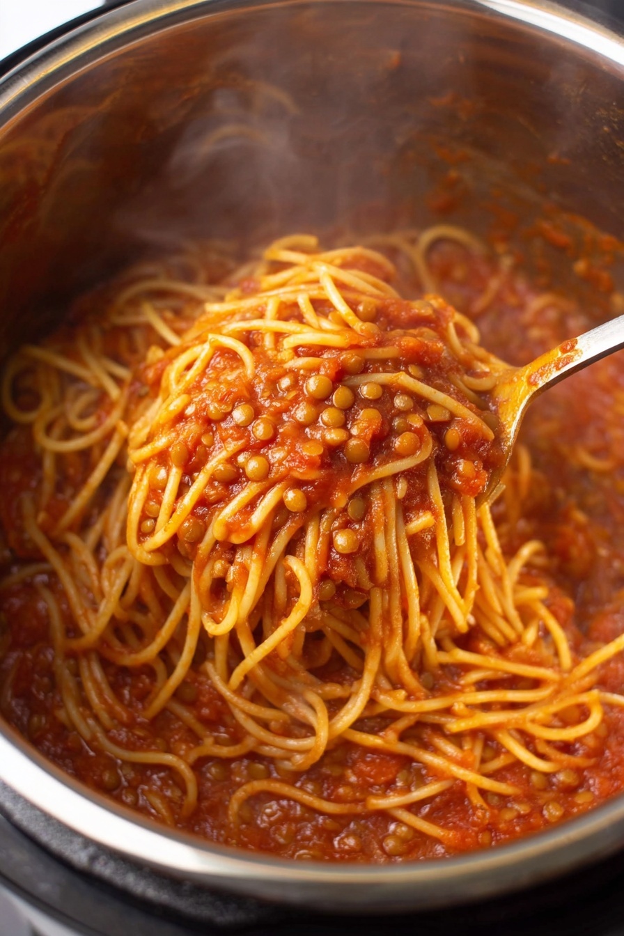 Instant Pot Lentil Spaghetti, healthy lentil pasta dinner, easy vegan pasta recipe, quick plant-based meals, one-pot lentil pasta - A close-up view inside a shiny stainless steel pot filled with spaghetti noodles mixed with small lentils in a rich red tomato sauce; the noodles have a soft yellow color and are coated well by the thick, slightly chunky sauce; a spoon is stirring the mixture, showing a tangled clump of noodles lifted above the rest with pieces of lentils visible around; steam rises gently from the hot dish. photo taken with an iphone --ar 2:3 --v 7