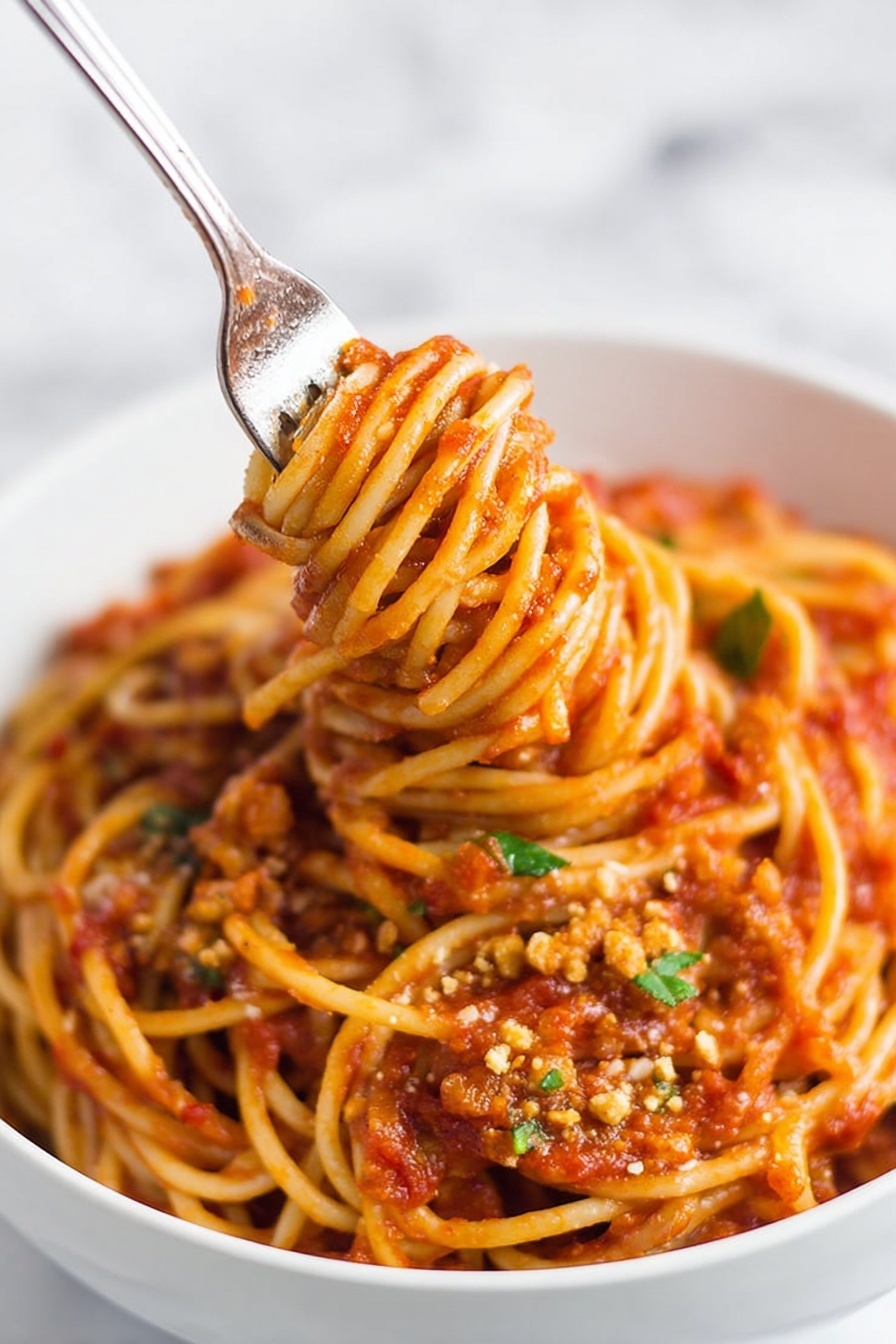Instant Pot Lentil Spaghetti, healthy lentil pasta dinner, easy vegan pasta recipe, quick plant-based meals, one-pot lentil pasta - A close-up of a white bowl filled with spaghetti noodles coated in rich red tomato sauce with visible small chunks and texture throughout. The spaghetti is thick and twisted around an inserted fork in the center, lifting the noodles upward, showing a dense mix of sauce and soft pasta strands. Small bits of green herbs and crumbled light brown topping are scattered on the surface, adding color contrast. The bowl is placed on a white marbled surface, and the image has a soft background with a focus on the noodles and sauce. photo taken with an iphone --ar 2:3 --v 7