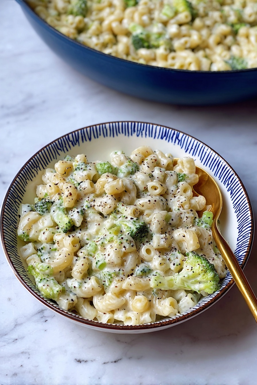 Creamy Broccoli Pasta with Cheesy Sauce, creamy broccoli pasta, cheesy pasta recipes, comforting broccoli pasta dish, easy cheesy pasta - A white bowl with a blue rim holds a creamy pasta dish made with small tube-shaped pasta mixed with small green broccoli pieces. The pasta is covered in a white, thick sauce, speckled throughout with black pepper. A gold spoon is placed on the right side of the bowl, partially resting in the pasta. Behind the bowl, there is a large blue pan filled with the same creamy pasta and broccoli mixture. The dish is set on a surface with a white marbled texture. photo taken with an iphone --ar 2:3 --v 7