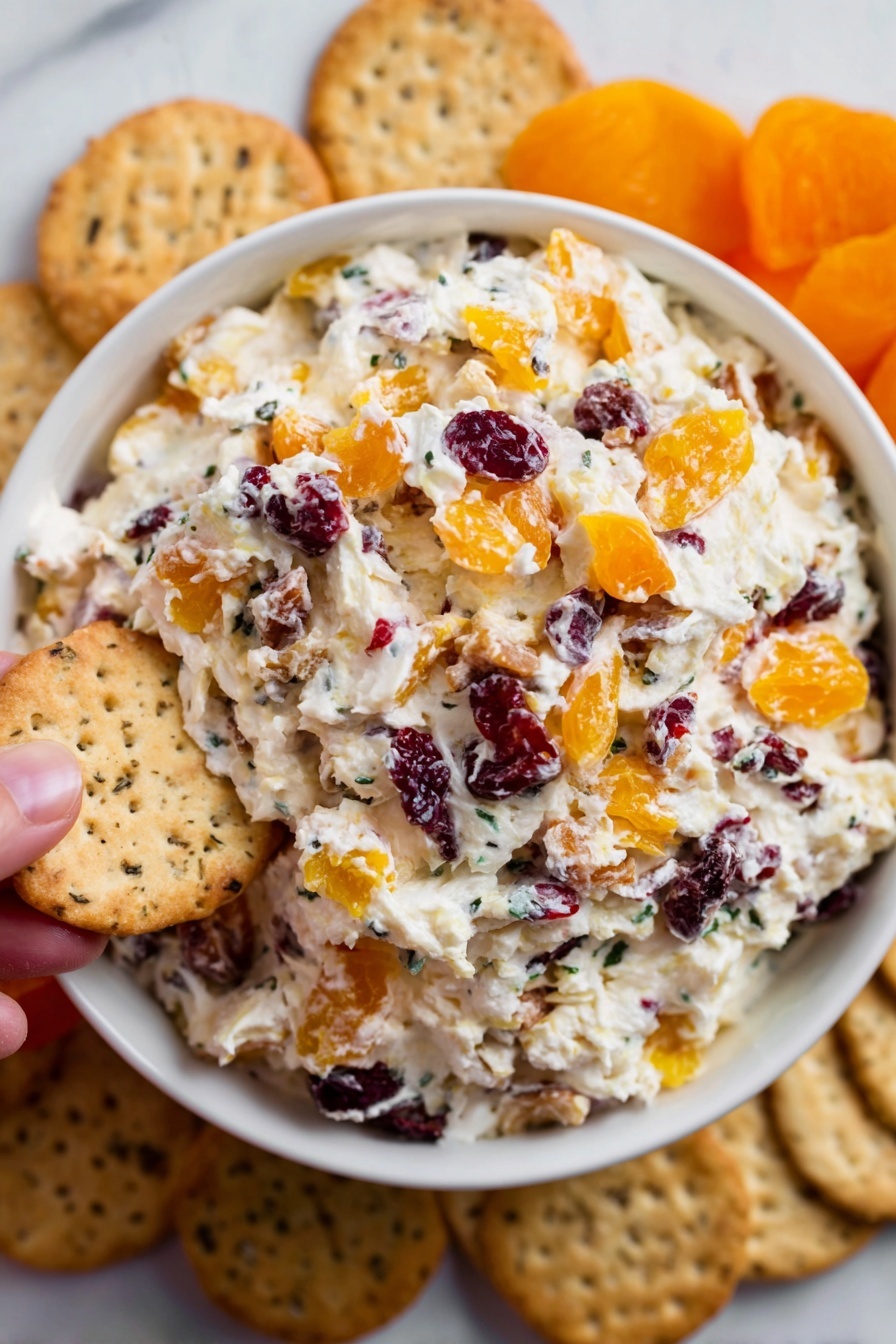 Cranberry Orange Cream Cheese Spread, quick fruit spreads, holiday appetizer ideas, creamy fruit spreads, easy brunch recipes - A white bowl is filled with a creamy mixture that has three layers of visible ingredients: the base is soft white cream cheese, mixed throughout with chunks of orange dried apricots and small dark red dried cranberries, creating a colorful and textured look. Around the bowl, there are beige crackers placed on a white marbled surface, some showing a speckled texture. A woman's hand is holding one of the crackers near the edge of the bowl. The scene is bright and clear, showing the fresh, inviting look of the snack. photo taken with an iphone --ar 2:3 --v 7