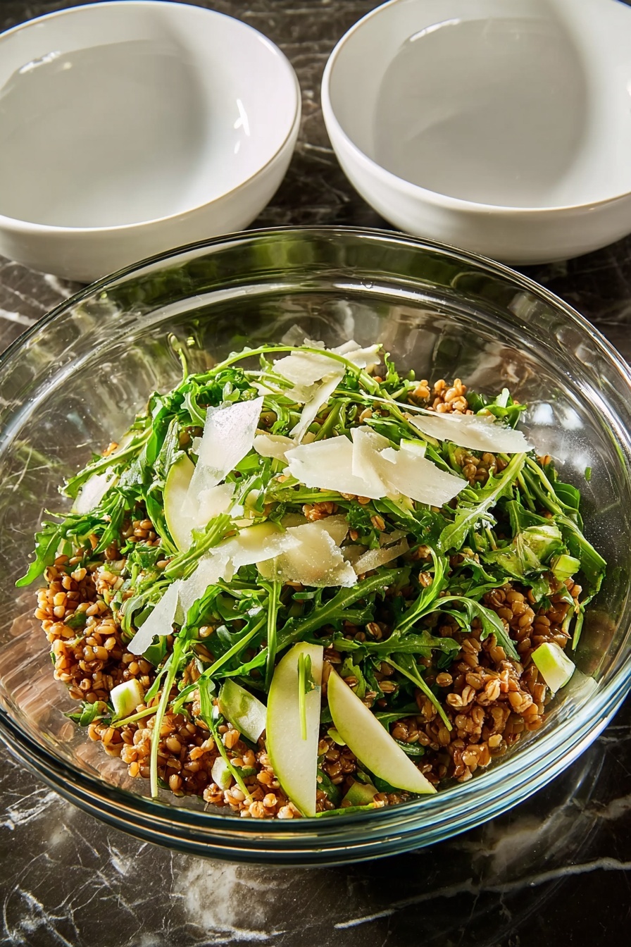 Farro Salad with Apple and Arugula, healthy farro salad, apple and arugula salad, easy grain salad, flavorful veggie salad - A large white round plate filled with a mixed salad that has several layers visible: the base layer is light brown cooked grains, mixed evenly with bright green arugula leaves scattered throughout. There are small, light green diced pieces of zucchini layered on top, along with thin, slightly browned slices of caramelized onions that add texture. Sprinkled lightly over the salad are thin shavings of light-colored cheese and small pieces of green herbs, giving a fresh look. The salad shows a mix of textures from soft grains to leafy greens and crunchy bits, all placed on a white marbled surface. photo taken with an iphone --ar 2:3 --v 7