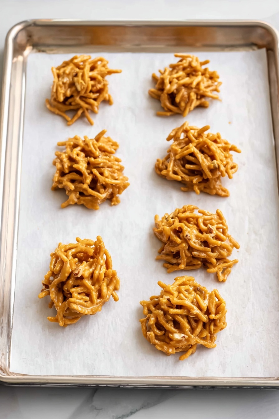 No Bake Haystack Cookies, easy no bake cookies, peanut butter chow mein cookies, no bake sweet treats, quick dessert recipes - There are six small, uneven clusters of golden brown candy made from thin, stick-like noodles mixed together with nuts, placed on white parchment paper that lines a silver baking tray. The candy clumps have a glossy, smooth texture from the coating, and they show some nut pieces poking out. The tray is placed on a white marbled surface. photo taken with an iphone --ar 2:3 --v 7