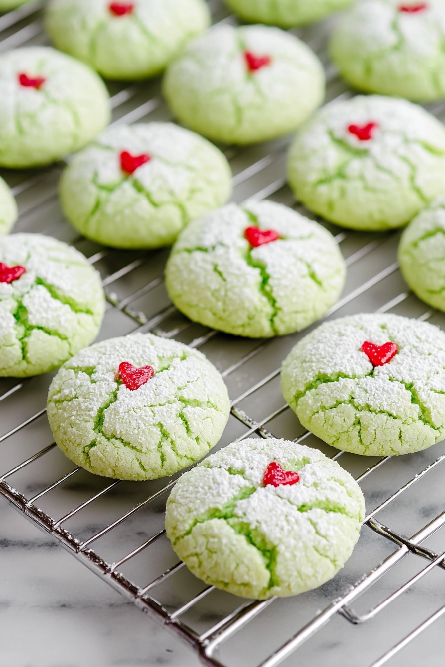 Green Peppermint Cake Mix Cookies, festive peppermint cookie recipe, easy holiday cookies, quick cake mix cookies, minty Christmas treats - The image shows rows of light green cookies on a silver cooling rack, each cookie round and cracked on top with a dusting of white powdered sugar. Each cookie has a small red heart-shaped decoration placed near the center. The cooling rack and cookies sit on a white marbled surface that is smooth and clean. The cookies appear soft and slightly puffy with a delicate texture. Photo taken with an iphone --ar 2:3 --v 7