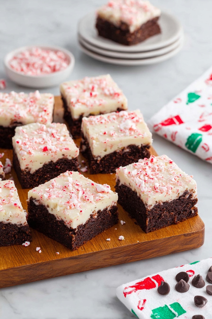 Peppermint Bark Brownie, Christmas brownie recipes, holiday peppermint desserts, fudge brownie with peppermint, festive chocolate treats - A wooden board holds eleven square brownies, each with two layers: a dark brown, dense textured base with visible chunks inside, topped by a creamy white layer sprinkled with crushed pink peppermint pieces giving a slightly rough texture. Three brownies are separated at the front, showing clear edges and layers. Behind the board, there is a small white stack of four plates with one brownie on top, and to the right, a white napkin with a pattern of small green and red chef hats rests on a white marbled surface. Scattered peppermint candies and a few chocolate chips decorate the bottom edge of the surface. Photo taken with an iphone --ar 2:3 --v 7