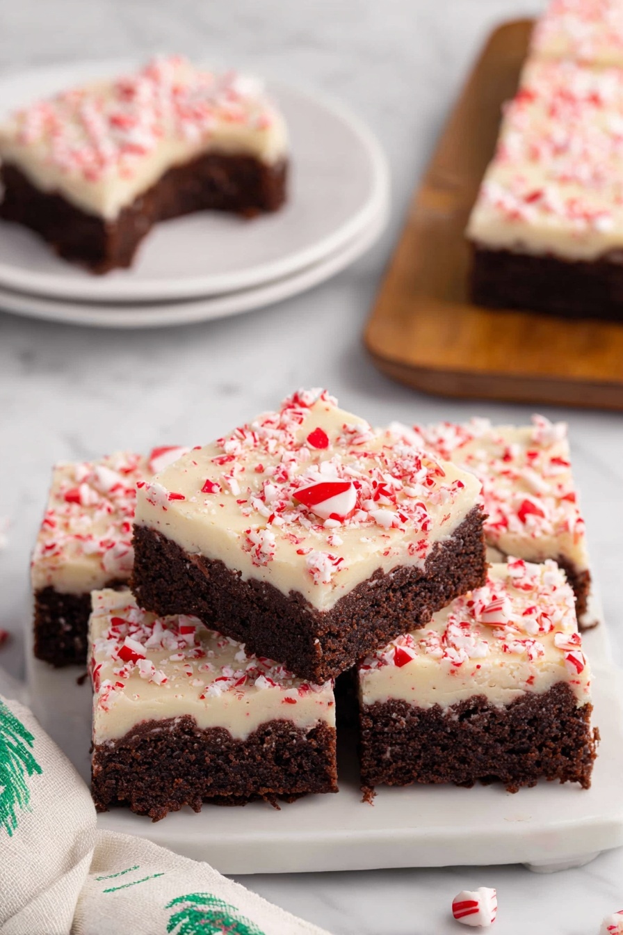 Peppermint Bark Brownie, Christmas brownie recipes, holiday peppermint desserts, fudge brownie with peppermint, festive chocolate treats - The image shows a stack of four square peppermint brownies on a white plate with a white marbled surface underneath. Each brownie has two layers: a thick, dark brown chocolate base with a moist, dense texture, and a creamy white topping sprinkled with crushed red peppermint bits that add a pop of color and texture. One brownie on top has a small bite taken out, revealing the rich chocolate layer inside. In the background, there is a similar white plate with another brownie on it, and a soft white cloth with green patterns is visible on the side. photo taken with an iphone --ar 2:3 --v 7