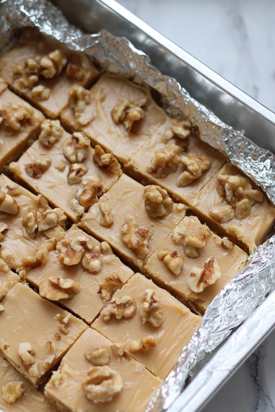 Maple Walnut Fudge, maple walnut fudge recipe, how to make maple walnut fudge, easy maple walnut fudge, homemade maple walnut fudge - The image shows a baking pan lined with crinkled aluminum foil holding a light beige fudge cut into small square pieces, topped with scattered walnut chunks. The fudge has a smooth but slightly cracked texture with the nuts unevenly spread on top. The pan rests on a white marbled surface. photo taken with an iphone --ar 2:3 --v 7