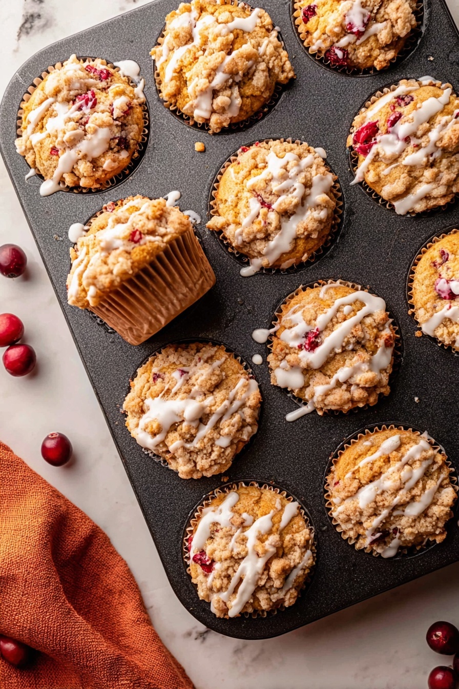 Cranberry Orange Muffins, Cranberry Orange Muffins Recipe, Easy Cranberry Muffins, Zesty Breakfast Muffins, Moist Cranberry Muffins - The image shows a dark muffin tin with nine muffins inside it, all golden brown with a crumbly topping that is light tan and unevenly spread over each muffin. Bright red berry pieces peek through the muffins, adding spots of color. White glaze is drizzled in thin lines over the top of each muffin, contrasting with the crumbly texture. One muffin is pulled slightly out of its spot showing the sides, which have a ridged paper liner of a light brown color. The muffin tin rests on a white marbled surface with a soft orange cloth partially visible in the bottom left corner and a few dark red whole berries scattered near the tin. photo taken with an iphone --ar 2:3 --v 7