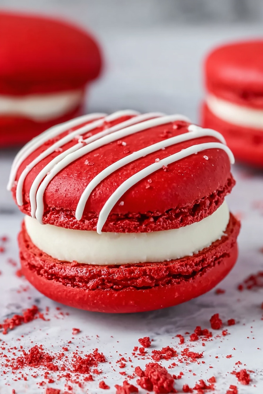 Red Velvet Macarons with Cream Cheese Filling, Red Velvet Macarons recipe, homemade red velvet macarons, cream cheese-filled macarons, festive red velvet desserts - The image shows a close-up of a bright red macaron with three visible layers: two smooth, round, glossy red shells with a slightly rough edge texture, and a thick white cream filling in the middle. The top shell is decorated with thin white icing lines running diagonally. The macaron rests on a white marbled surface with some white drizzle and red crumbs around it, adding detail and texture. In the background, there is a blurred second red macaron. Photo taken with an iphone --ar 2:3 --v 7