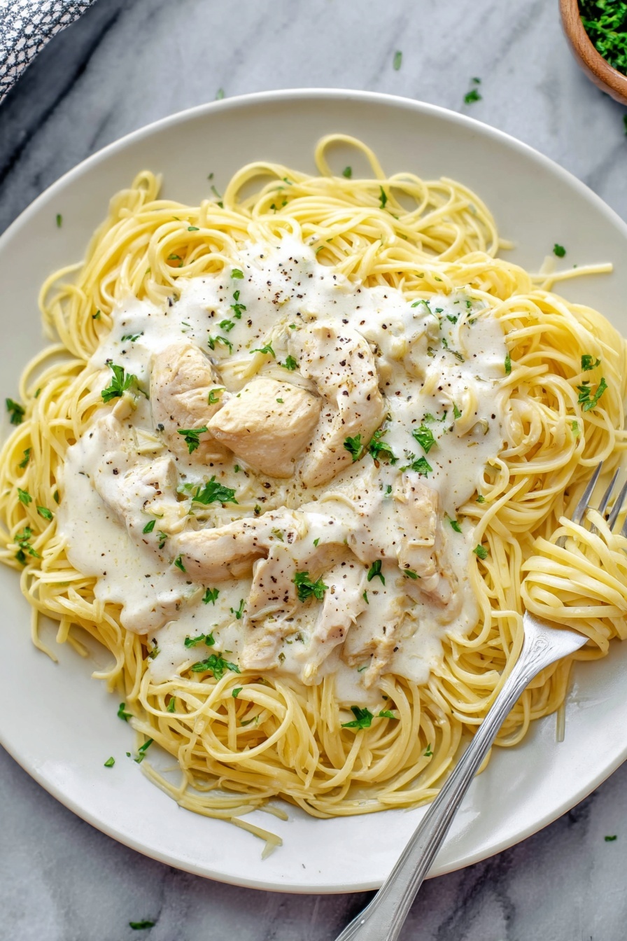 Creamy Crock Pot Angel Chicken, easy crock pot chicken recipes, rich creamy chicken dinner, foolproof slow cooker chicken, comforting crock pot chicken dish - A white plate holds a dish of thin yellow spaghetti noodles arranged loosely in a circle. On top of the noodles are several pieces of light cream chicken, covered with a thick white creamy sauce that has small bits of herbs and black pepper sprinkled over it. Fresh green parsley leaves are scattered on top of the sauce for a touch of color. A silver fork rests on the right side of the plate, twirling some noodles with sauce. The plate sits on a white marbled surface. Photo taken with an iphone --ar 2:3 --v 7