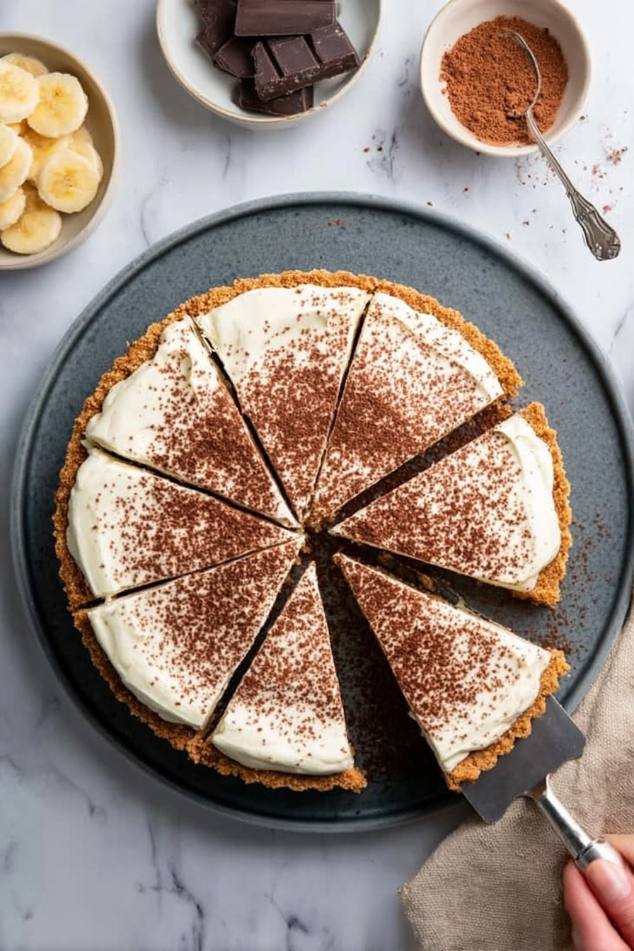 Banoffee Pie with Caramel and Bananas, how to make Banoffee Pie, easy Banoffee dessert, homemade Banoffee Pie, classic Banoffee pie recipe - The image shows a round tart with a crumbly, golden brown crust as the bottom layer. On top is a thick, white creamy layer spread evenly and sprinkled with fine brown cocoa powder covering most of the surface. The tart is sliced into six pieces with one slice being lifted by a metallic pie server. The tart is placed on a round dark gray plate, all on a white marbled surface. Nearby, there is a small white bowl with light yellow banana slices and another white bowl with brown cocoa powder and two dark chocolate pieces. A woman's hand holds the pie server under the lifted slice. Photo taken with an iphone --ar 2:3 --v 7