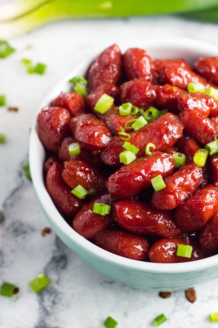 Crockpot BBQ Little Smokies, easy appetizer recipes, slow cooker party snacks, game day finger foods, crowd-pleasing smoky appetizers - The image shows a white bowl filled with small, shiny red sausage-like pieces covered in a glossy sauce, giving them a sticky and rich appearance. Bright green chopped scallions are scattered evenly over the sausages, adding a fresh contrast in color. The bowl sits on a white marbled surface with a few scattered scallions around it, creating a clean and simple background. The focus is close up and clear, highlighting the texture and color of the sausages and scallions. photo taken with an iphone --ar 2:3 --v 7