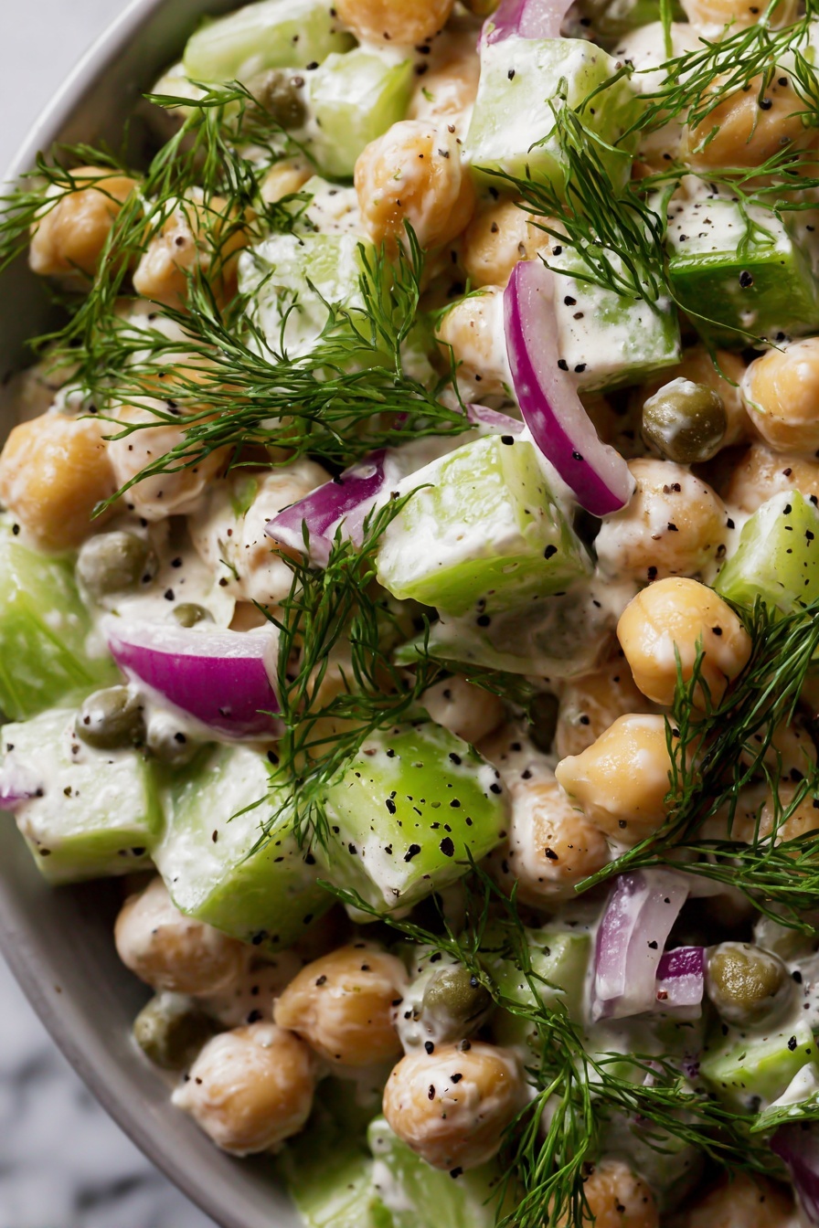 Healthy Tuna Chickpea Salad, healthy tuna chickpea salad, nutritious tuna salad, light tuna and chickpea dish, protein-packed salad - The image shows a close-up of a salad with several layers and colors. The main elements are round chickpeas that are beige and smooth, mixed with small cubes of light green celery and purplish red onion. There are round, green capers scattered throughout. Fresh green dill sprigs are placed on top, adding a feathery texture. The salad is coated with a creamy, white dressing that has small black seeds mixed in. The entire dish sits against a white marbled surface in soft light. photo taken with an iphone --ar 2:3 --v 7