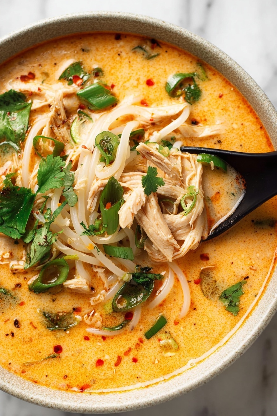 Thai Chicken Curry Soup, Thai chicken coconut soup, easy Thai curry soup, spicy chicken coconut soup, quick Thai soup recipes - A white bowl filled with creamy orange broth covering soft white noodle strands arranged in the center and slightly tangled. On top, several shredded pale chicken strips are spread out, mixed with small dark green herb pieces and chopped light green onions. A bright green spinach leaf rests near the middle, surrounded by fresh cilantro leaves scattered around the edge. A wedge of lime with visible texture sits on the left side, partially submerged in the broth. The bowl is placed on a white marbled surface. photo taken with an iphone --ar 2:3 --v 7