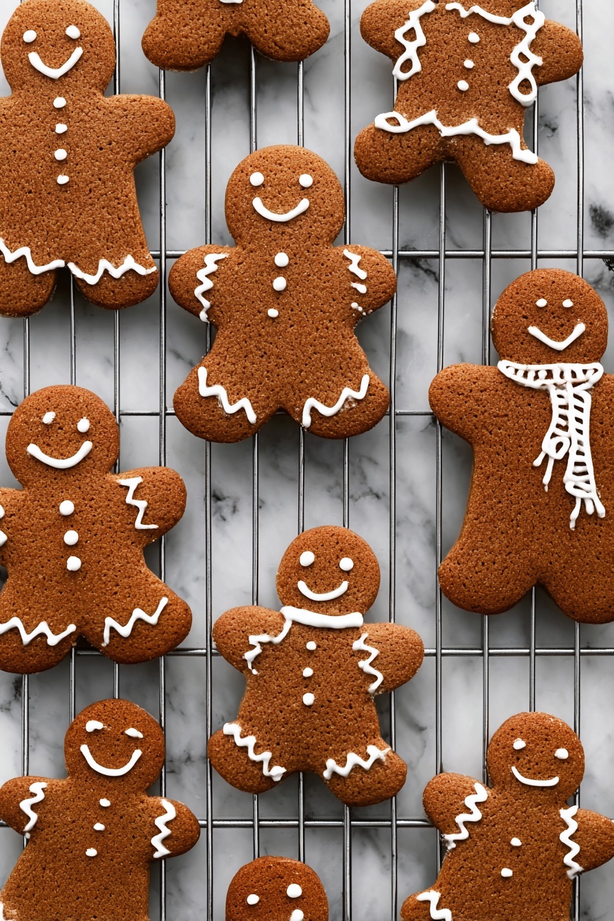 Gingerbread Men Cookies, Gingerbread Cookies Recipe, Festive Gingerbread Cookies, Homemade Gingerbread Men, Holiday Cookie Recipes - The image shows several gingerbread cookies shaped like people, placed on a metal cooling rack over a white marbled texture. Each cookie is a warm brown color with a rough texture and decorated with white icing in different patterns: some have smiles, eyes, buttons, and zigzag designs on arms and legs, while others have scarves or simple outlines on the edges. The cookies are arranged in various sizes and angles across the cooling rack, creating a cozy and festive feel. photo taken with an iphone --ar 2:3 --v 7