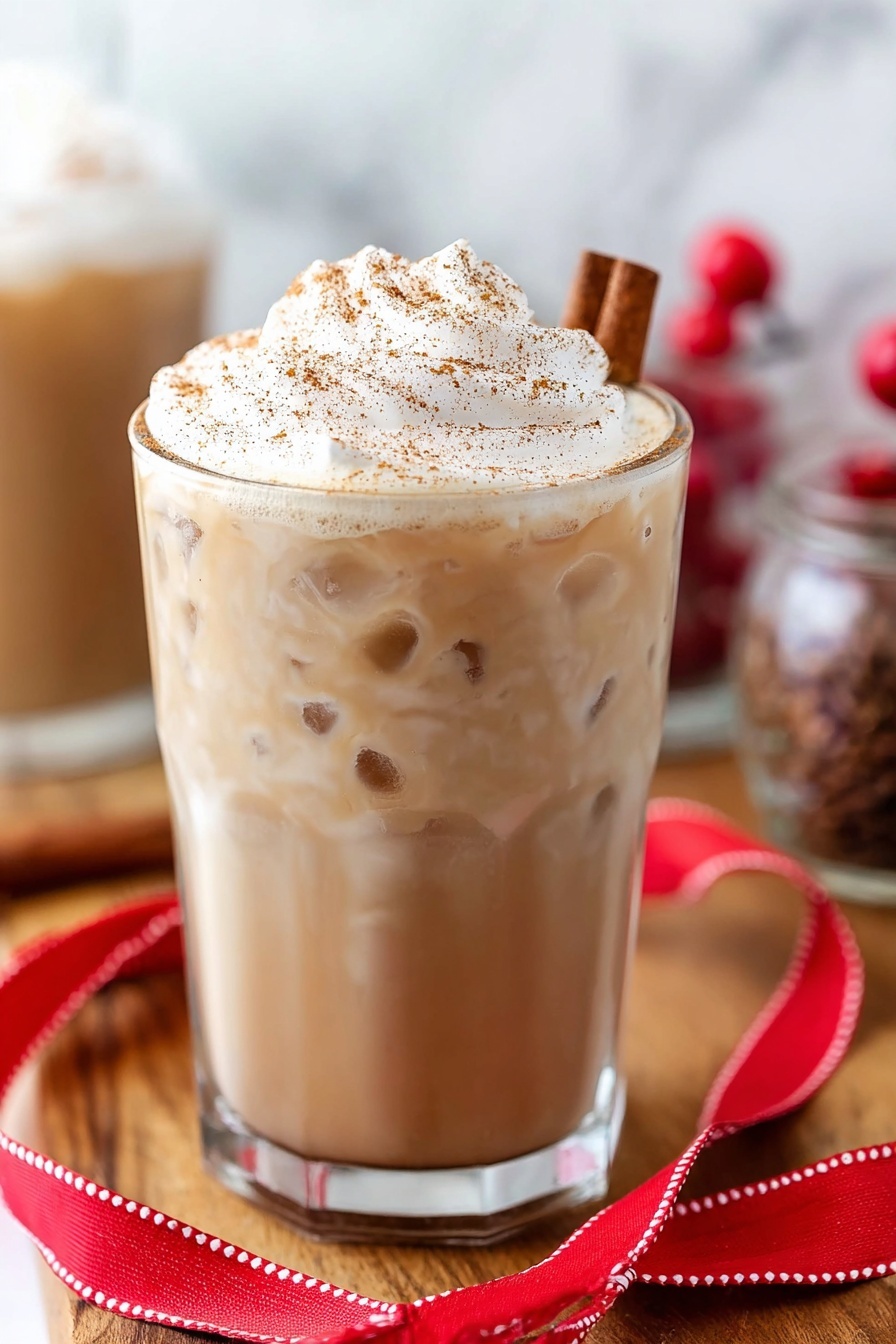 Eggnog White Russian Cocktail, festive holiday cocktails, creamy whiskey drinks, easy holiday cocktail recipes, eggnog cocktails - A clear glass is filled almost to the top with a creamy light brown drink. On top, there is a tall, fluffy layer of white whipped cream sprinkled with brown spice. A cinnamon stick is placed in the whipped cream on the right side. The glass sits on a wooden surface, with a folded red ribbon with white stitching lying in front of it. There are some small red berries blurred in the background along with other glass containers. The background has a white marbled texture. photo taken with an iphone --ar 2:3 --v 7