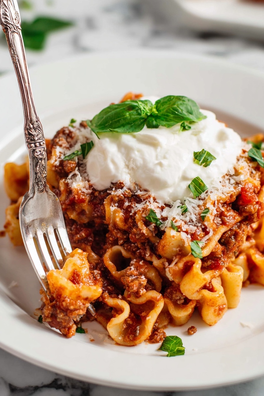 Quick Homemade Lasagna Hamburger Helper, easy lasagna skillet dinner, one-pot lasagna recipe, quick ground beef dinner, cheesy lasagna pasta - In the image, there is a white plate on a white marbled surface with pasta mixed with a thick red-brown meat sauce spread across it. On top of the pasta, there is a dollop of white cream with a small green basil leaf placed on it. Thin green basil strips and a sprinkle of shredded white cheese are scattered around the pasta on the plate. A silver fork rests on the edge of the plate. Behind this plate, a larger white bowl filled with more of the pasta dish sits on a light cloth, garnished with a whole basil leaf in the center. To the upper right, part of another white plate with the same pasta dish is visible. Small red cherry tomatoes are scattered at the bottom right corner on the white marbled surface, and a small shaker with red pepper flakes is placed to the left of the main plate. The scene is cozy and inviting, all set on the clean white marble background. photo taken with an iphone --ar 2:3 --v 7