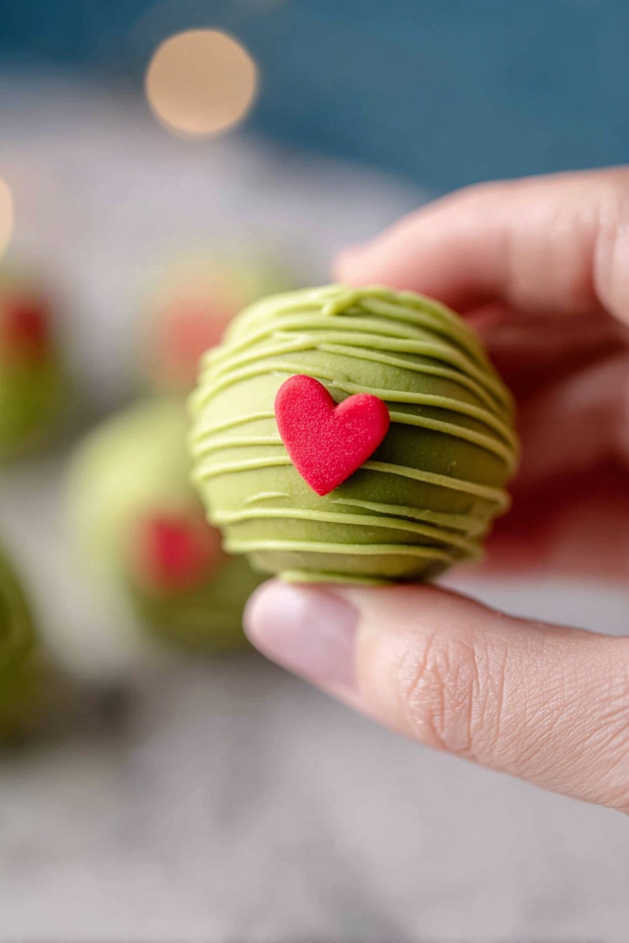 Grinch Oreo Balls with Red Velvet Oreos, Festive Christmas Oreo Bites, Holiday Treats with Red Velvet Oreos, Christmas Dessert Balls, Easy Festive Oreo Snacks - This image shows a white bowl filled with round green balls that have smooth surfaces and are drizzled with thin lines of a lighter green shade. Each ball is topped with a small red heart-shaped decoration. The bowl rests on a matching white plate, both placed on a white marbled surface. Around the plate are a few more of the red heart shapes scattered softly. The background is softly blurred with soft warm lights and out-of-focus colorful holiday decorations visible. Photo taken with an iphone --ar 2:3 --v 7