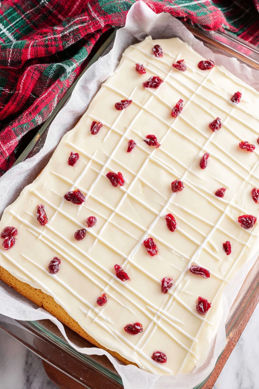 Cranberry White Chocolate Bliss Bars, cranberry white chocolate bars, festive dessert bars, easy holiday treats, moist white chocolate bars - A rectangular layered dessert lies on white parchment paper inside a glass baking dish on a white marbled surface. The bottom layer is golden brown, thin, and even. On top is a thick, smooth white frosting layer covering the entire surface. Thin white icing lines are drizzled across the frosting, forming a light grid pattern. Scattered over the top are small dried red fruit pieces, evenly spaced, adding bright red spots to the white frosting. The cloth nearby has green, red, and dark colors in a check pattern. photo taken with an iphone --ar 2:3 --v 7