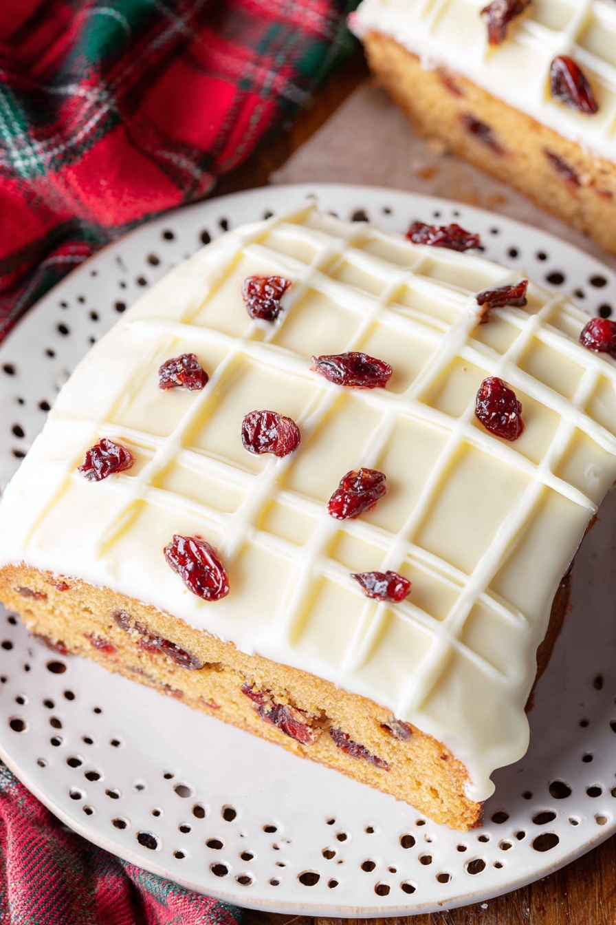 Cranberry White Chocolate Bliss Bars, cranberry white chocolate bars, festive dessert bars, easy holiday treats, moist white chocolate bars - A single slice of cake is shown on a white decorative plate with small cut-out patterns. The cake has two clear layers: a dense golden-brown base with visible small dark red dried fruit pieces inside, and a thick smooth white icing layer on top, decorated with a white grid pattern and scattered dark red dried fruit pieces. The plate rests on a wooden surface with a blurred part of the whole cake and a red and green fabric in the background. Photo taken with an iphone --ar 2:3 --v 7
