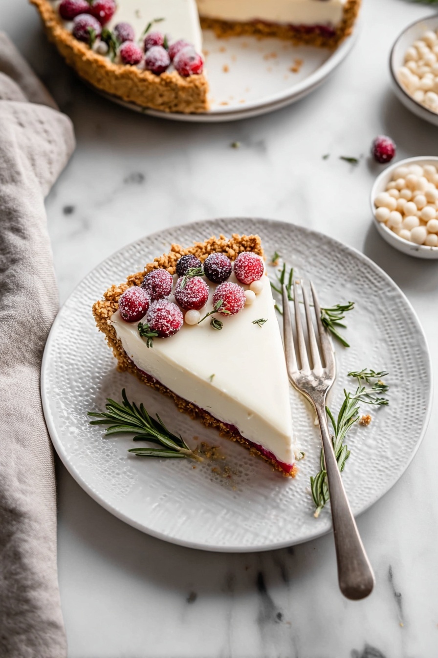 Cranberry White Chocolate Tart, cranberry white chocolate dessert, holiday tart recipes, easy cranberry tart, white chocolate cranberry pie - The image shows a slice of pie on a white plate with a textured rim, placed on a white marbled surface. The pie has three visible layers: a golden brown crumbly crust, a thin red layer in the middle, and a thick, smooth white topping. The slice is decorated on one side with sugared red cranberries, a few small white drops, and sprigs of fresh green rosemary. A silver fork rests on the right side of the plate. Nearby, there is a whole pie with a similar topping and decoration, and a bowl filled with small white drops. A light gray cloth is placed towards the bottom left corner of the image. Photo taken with an iphone --ar 2:3 --v 7