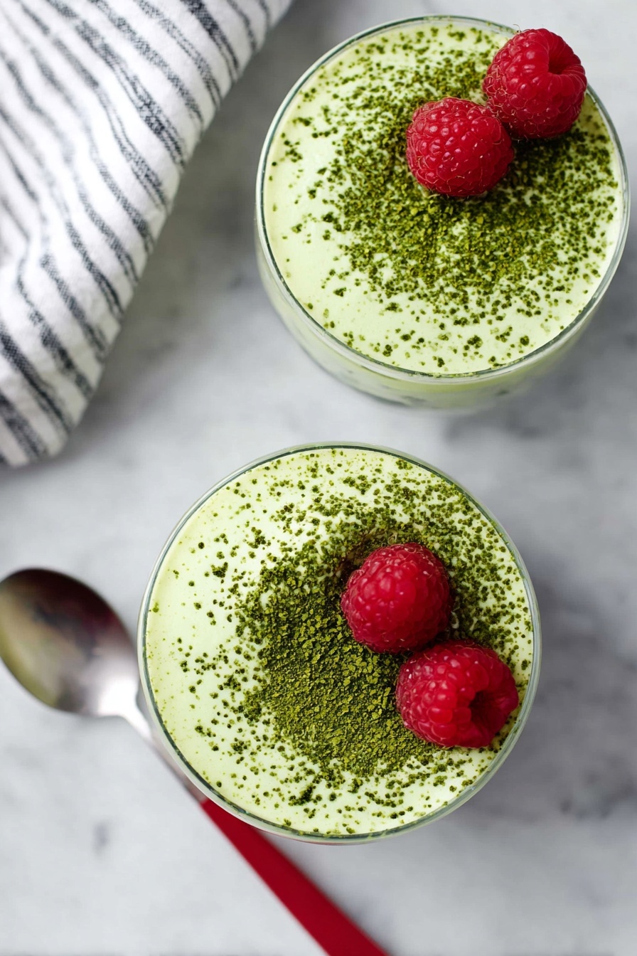 Matcha Protein Chia Pudding, healthy matcha chia pudding, protein-packed breakfast recipes, quick nutritious snacks, vegan chia pudding ideas - Two round glasses filled with a creamy light green mousse topped with a dusting of darker green powder evenly spread on the surface. Each mousse has two bright red raspberries placed near the top edge. The glasses sit on a white marbled surface. A spoon with a red handle is placed next to the bottom glass. Part of a white and black striped cloth is visible on the top left corner. Photo taken with an iphone --ar 2:3 --v 7
