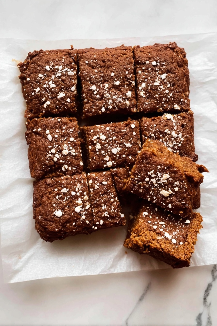 Healthy Apple Oatmeal Cake, healthy breakfast cake, gluten-free apple cake, wholesome oatmeal cake, easy apple oatmeal dessert - The dish is a rectangular brown baked cake cut into nine pieces with a rough texture and a slightly cracked top. The cake is sprinkled with white oat flakes spread unevenly over the surface. It rests on white parchment paper placed on a white marbled surface. One piece in the top right corner is separated from the rest, showing the inside of the cake, which is soft and dense with a uniform brown color. photo taken with an iphone --ar 2:3 --v 7