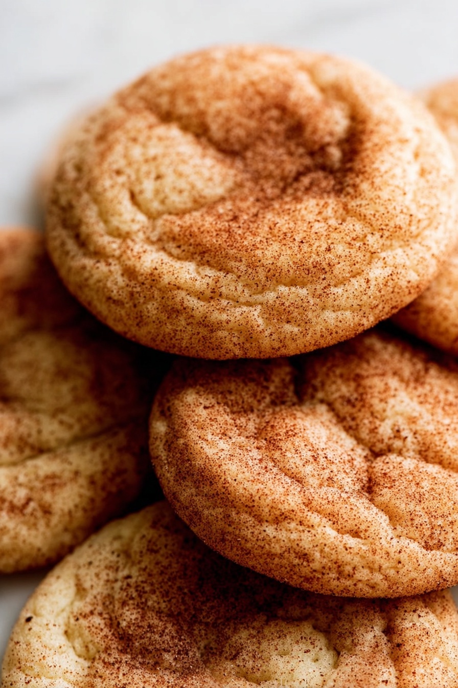 Brown Butter Snickerdoodles, cinnamon sugar cookies, easy brown butter cookies, soft chewy snickerdoodles, homemade cinnamon cookies - A close-up view of a pile of soft, round cookies stacked unevenly on a white marbled surface. Each cookie has a light golden base with a sprinkled coating of cinnamon sugar, giving them a warm brown speckled texture. The cookies show gentle cracks and slight indentations on the surface, highlighting their soft and chewy texture. The focus is tight, capturing the detailed cinnamon dusting and the uneven round shapes of the cookies. photo taken with an iphone --ar 2:3 --v 7